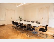 Moderner Besprechungsraum mit einem langen weißen Tisch und zwölf schwarzen Stühlen auf einem hellen Parkettboden. Die Wände sind cremefarben, mit dezenten Zierleisten, zwei Türen und einem Sideboard mit einer Pflanze darauf.