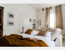 Helles Wohnzimmer mit einem aufklappbaren Sofa als Bett, einer senfgelben Decke und goldfarbenen Kissen. Im Hintergrund ein Esstisch mit vier Stühlen vor bodenlangen beige Vorhängen, dekoriert mit Wandbildern, hängenden Lampen und einem kleinen Spiegel.