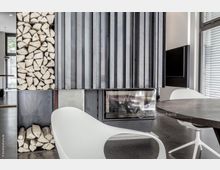 Moderner Innenraum mit einem Kamin, der teilweise von vertikalen Metallpaneelen verkleidet ist. Links befindet sich ein Holzlager, rechts moderne weiße Stühle und ein runder Tisch vor einem großen Fenster mit Jalousien.