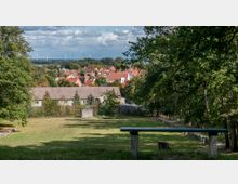 Eine ländliche Szenerie mit Blick auf ein Dorf mit roten Ziegeldächern und einer Backsteinkirche im Zentrum, umgeben von grünen Wiesen, Bäumen und Windrädern am Horizont. Im Vordergrund befindet sich eine grüne Holzbank und eine offene Rasenfläche, eingerahmt von Bäumen.