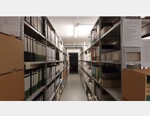 Ein Archivraum mit langen, parallelen Regalreihen, gefüllt mit Ordnern, Akten und Archivboxen. Der Raum ist schlicht beleuchtet, mit einer sichtbaren Deckenleuchte und einem Durchgang am Ende des Gangs.
