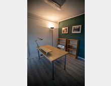 Ein einfacher Büroraum mit einem Holztisch und einer weißen Kunststoffstuhl, flankiert von zwei leeren Holzregalen. Die Wände sind grün und weiß gestrichen, mit zwei gerahmten Bildern an der grünen Wand und einer Stehlampe in einer Ecke.