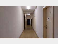 Ein langer, schlichter Flur mit weißen Wänden und einem Boden aus glänzendem Beton. Rechts und links befinden sich mehrere Türen, und an der Decke sind Leuchtstofflampen angebracht.