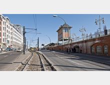 Straßenszene mit Straßenbahnschienen in der Mitte, flankiert von Fahrbahnen und Gehwegen. Auf der rechten Seite befindet sich eine historische Backsteinmauer mit gewölbten Fenstern und einem erhöhten Bahnkontrollturm, während links moderne Gebäude und parkende Fahrräder zu sehen sind.