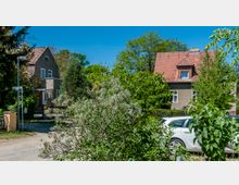 Zwei Einfamilienhäuser mit roten Satteldächern stehen in einer grünen, von Bäumen und Sträuchern umgebenen Wohngegend. Im Vordergrund ist ein geparkter weißer PKW und ein Straßenschild sichtbar.