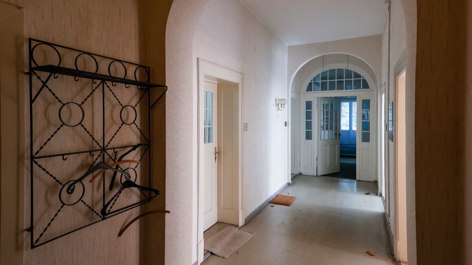 Langer, schmaler Flur in einem älteren Gebäude mit hohen Rundbögen, hellen Wänden und grauem Boden. Links hängt eine schwarze Metallgarderobe, und am Ende führen verglaste Doppeltüren unter einem Rundbogen in einen weiteren Raum.