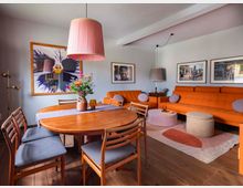 Wohnzimmer-Interieur mit Holzboden, einer orangefarbenen Polstercouch, einem runden Holztisch mit Stühlen und einer großen, rosa Hängelampe. An den Wänden hängen gerahmte Kunstwerke und Fotografien, während bunte Dekorationen wie ein Blumenstrauß und kleine Beistelltische den Raum ergänzen.