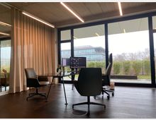 Modernes Büro mit Glasfront, die den Blick auf einen Garten mit Hecke und ein modernes Gebäude im Hintergrund freigibt. Im Vordergrund stehen ein Glastisch mit zwei Bürostühlen, zwei Computermonitore und eine Vase mit Blumen; der Raum hat eine helle Vorhangwand und eine Decke mit Betonoptik.