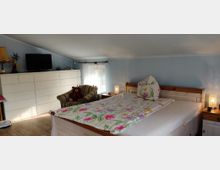 Ein gemütliches Schlafzimmer mit hellblauen Wänden und Dachschräge. Ein Holzbett mit bunter Bettwäsche steht rechts, daneben zwei Nachttischlampen; links ein weißer, langer Schrank mit aufgesetztem Fernseher und ein gestreifter Sessel mit einem Stofftier.