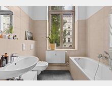 Helles Badezimmer mit cremefarbenen Fliesen, einer Badewanne, einem Waschbecken, einer Toilette und einer großen Fensterfront. Auf der Fensterbank steht eine Zimmerpflanze und im Hintergrund ist ein Gebäude zu sehen.
