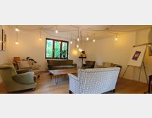 Ein gemütlicher Raum mit einer Sitzgruppe aus Polstersesseln und Sofas, die um einen Holztisch arrangiert sind. An der Decke hängen freiliegende Glühbirnen, ein Fenster mit Blick auf grüne Bäume, eine Stehlampe und ein Flipchart befinden sich im Hintergrund.