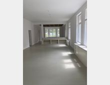 Ein leerer, heller Raum mit hellgrauem Boden, weißen Wänden und großen Fenstern, die Tageslicht hereinlassen. Im Hintergrund befindet sich eine gemauerte Durchreiche mit Blick auf weitere Fenster und dichten grünen Bewuchs außerhalb.