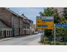 Straßenszene in einer kleinen Stadt mit mehreren Wohnhäusern entlang der Straße. Im Vordergrund stehen gelbe Verkehrsschilder mit Wegweisern zu umliegenden Städten wie Trebbin, München und Bad Belzig, sowie ein Straßenschild mit der Aufschrift „Berliner Straße“.