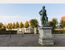 Statue von Kronprinz Friedrich auf einem steinernen Sockel, umgeben von einem gepflasterten Platz. Im Hintergrund befinden sich eine Toranlage, gepflegte Hecken, eine Baumreihe und ein historisches Gebäude mit roten Ziegeldächern.