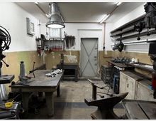 Werkstatt mit industriellem Charakter, ausgestattet mit einem großen Arbeitstisch in der Mitte, Werkzeugen an der Wand, mehreren Maschinen und Werkbänken entlang der Seiten. Im Hintergrund befindet sich eine Metalltür, und die Beleuchtung erfolgt über Deckenlampen und eine hängende Industrieleuchte.