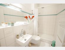 Modernes Badezimmer mit weißen Fliesen, einer bodengleichen Dusche mit Duschvorhang, einem kleinen Waschbecken und einer wandhängenden Toilette. Dekorative Elemente umfassen kleine Behälter, gefaltete Handtücher und Vasen mit roten Trockenblumen.