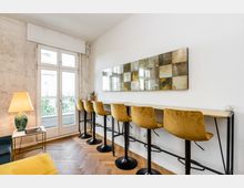 Heller Innenraum mit langem Hochtisch entlang einer weißen Wand, davor mehrere Barhocker mit senfgelben Polstersitzen und einer großen rechteckigen Wandkunst aus spiegelnden Kacheln. Links führen hohe Glastüren auf einen Balkon; der Raum hat Fischgrätparkett, eine Stehlampe und eine teils unverputzte Wandfläche.