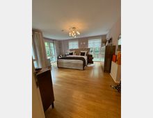 Großes Schlafzimmer mit Holzboden, einem großen Bett in der Mitte mit gepolstertem Kopfteil, dekorativen Kissen und Nachttischen. Helle Wände, große Fenster mit Vorhängen und weißen Rollläden sowie eine moderne Deckenlampe definieren den Raum.