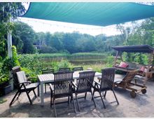 Terrasse mit Holzstühlen und einem Tisch auf einem gepflasterten Boden, umgeben von Pflanzen und mit Blick auf einen Teich und dahinterliegenden Wald. Ein grünes Sonnensegel und eine Holzwiege mit Sitzpolstern ergänzen die Szenerie.