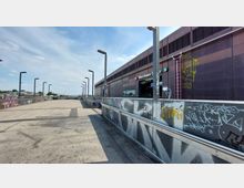 Eine erhöhte Fußgängerbrücke mit Laternen, deren Geländer mit Graffiti bedeckt sind, führt zu einem modernen, dunkelbraunen Bahnhofsgebäude mit dem Schild "Warschauer Straße". Im Hintergrund sind Gebäude und ein klarer Himmel zu sehen.