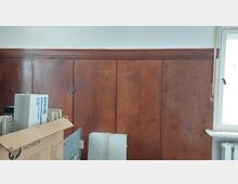 Ein Raum mit einer Wandverkleidung aus dunkelbraunem Holz, teilweise mit Rissen in der Lackierung. Rechts befindet sich ein weißer Fensterrahmen mit einem Heizkörper darunter, und im Vordergrund stehen Kartons sowie Elektronikgeräte.