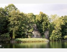 Historisches Gebäude aus Naturstein mit Satteldach, teilweise von hohen, dicht bewachsenen Bäumen verdeckt, am Ufer eines ruhigen Sees mit Schilfgürtel im Vordergrund.