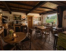 Gemütlicher Gastraum eines rustikalen Cafés mit Holzbalkendecke, Ziegelboden und mehreren Holztischen und -stühlen, teils mit Kerzen und Blumen dekoriert. An den weiß verputzten Wänden hängen Bilder und Lampen; ein großes Fenster mit Gitter blickt ins Grüne.