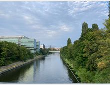 Ein kanalartiger Fluss, der von dicht bewachsenen Bäumen auf der rechten und von modernen Glasgebäuden auf der linken Seite gesäumt ist, mit einer Brücke im Hintergrund unter einem leicht bewölkten Himmel.