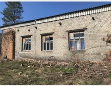 Eine verlassene Backsteinmauer mit drei großen Fenstern, von denen einige beschädigt oder mit Holz verschlossen sind. Im Vordergrund befinden sich Überreste von Ziegelsteinen und trockenes Gras, während im Hintergrund ein blauer Himmel sichtbar ist.
