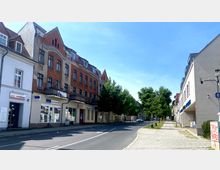 Straßenszene mit einer Reihe von mehrstöckigen Gebäuden, darunter ein Backsteinhaus mit Erkern und Balkonen, sowie Geschäften im Erdgeschoss. Auf der rechten Seite verläuft ein Gehweg mit gepflastertem Boden neben einer Fahrbahn, die von Bäumen gesäumt ist.