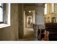 Ein alter, verlassener Kirchenraum mit abblätternder Wandfarbe, Holzbänken auf der rechten Seite und hohen Rundbogenfenstern, die gedämpftes Licht hereinlassen. Im Hintergrund ist ein schmaler Gang mit einer Tür zu sehen.
