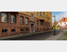 Straßenszene mit mehrstöckigen Wohnhäusern aus Backstein und Putzfassaden, darunter ein gelb gestrichenes Obergeschoss. Gepflasterter Gehweg und asphaltierte Straße mit Straßenlaternen führen entlang der Gebäude. Im Hintergrund steht ein weiteres Gebäude mit rotem Ziegeldach.