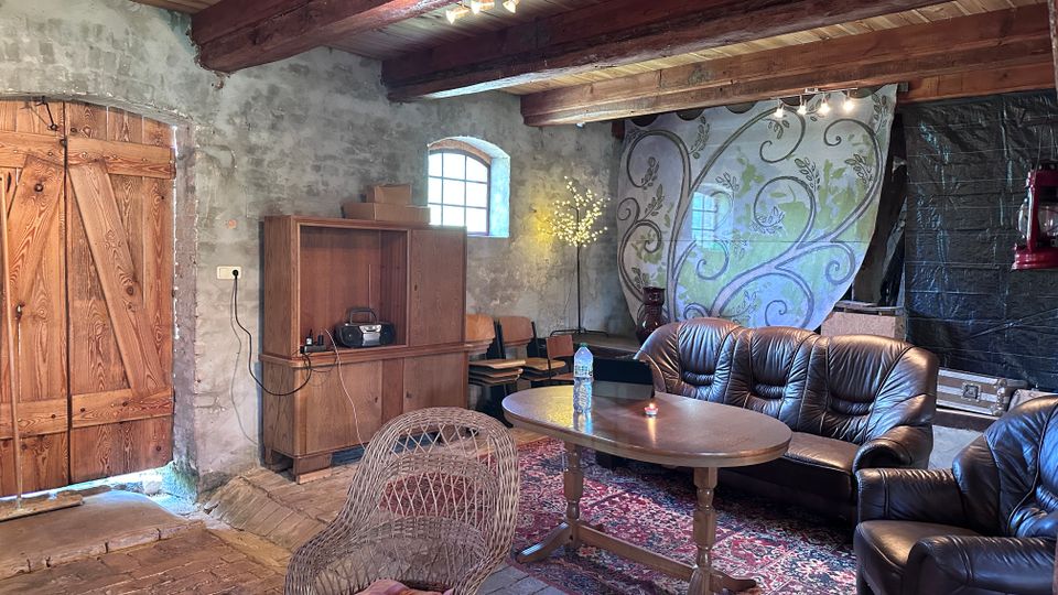 Rustikaler Innenraum mit groben Steinwänden, sichtbaren Holzbalken und einem großen Holztor neben einem kleinen Rundbogenfenster. Im Vordergrund stehen Ledersofas um einen ovalen Holztisch auf einem Teppich; im Hintergrund sind ein Holzschrank, gestapelte Stühle und eine dekorative bemalte Wand zu sehen.