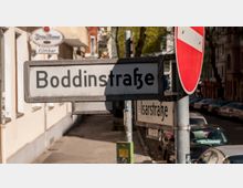Straßenschild mit der Bezeichnung „Boddinstraße“ im Vordergrund, darunter ein weiteres Schild mit der Aufschrift „Isarstraße“. Rechts daneben ein rot-weißes Verkehrsschild „Durchfahrt verboten“. Im Hintergrund sind geparkte Autos, eine Hausfassade und Bäume zu erkennen.