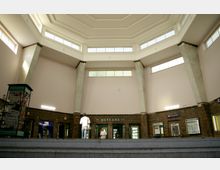 Innenaufnahme eines Bahnhofsgebäudes mit einer hohen, oktagonalen Decke und großen Fenstern, die Tageslicht einlassen. Im unteren Bereich sind Klinkerwände mit mehreren Türen, Schaltern für Fahrkarten und Informationsständen, sowie ein Schild mit der Aufschrift „Ausgang“ sichtbar.