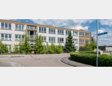 Außenansicht eines dreistöckigen, rechteckigen Gebäudes mit vielen gleichmäßigen Fenstern, umliegenden Bäumen und einem Gehweg im Vordergrund. An der Straßenecke ist ein Straßenschild mit der Aufschrift „Norderstedter Str.“ zu sehen.