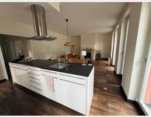 Moderne Wohnung mit offenem Küchen- und Wohnbereich. Im Vordergrund eine weiße Kücheninsel mit dunkler Arbeitsplatte und eingebautem Waschbecken, im Hintergrund ein Essbereich, ein heller Kamin und bodentiefe Fenster auf der rechten Seite.