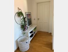 Schmaler Flur mit Parkettboden und einer weißen Tür am Ende, links ein weißes Regal mit offenen Fächern und dekorativen Gegenständen, darüber ein gerahmtes Bild sowie eine hängende Pflanze im Ring, unten links ein gestreifter Wäschekorb.