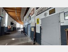 Innenansicht eines historischen Werkstattraumes mit weißen Wänden, teilweise dunkel gestrichenem unteren Bereich und hölzerner Decke. Entlang der Wände sind zahlreiche alte Schilder, Werkzeuge und Anschauungstafeln angebracht; im Hintergrund befinden sich große Fenster und weiteres altes Equipment, darunter eine rote Waage.