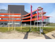 Modernes Gebäude mit flachen, roten, geschwungenen Dachelementen und schwarzer Fassade. Im Vordergrund stehen junge Bäume, ein Weg und grüne Rasenflächen, daneben mehrere Fahnenmasten mit lilafarbenen Bannern.