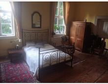 Schlafzimmer mit einem schmiedeeisernen Bettgestell, einem Nachttisch und einem hölzernen Kleiderschrank. Der Raum hat Holzboden, gelb gestrichene Wände, große Fenster mit Vorhängen und ist mit Teppichen dekoriert.