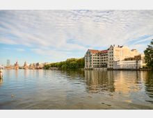 Blick über einen breiten Fluss auf ein Ufer mit einem großen, mehrstöckigen Gebäudeensemble mit Satteldächern und vielen Fenstern, teils mit Gerüst und Verkleidung. Im Hintergrund ist eine markante Brücke mit Türmen sowie weitere Stadtbebauung und eine Baumreihe entlang des Ufers zu sehen.