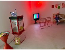 Ein schlicht eingerichteter Raum mit roten Lichtakzenten, einer Popcornmaschine, zwei Metallstühlen, einem alten Fernseher mit Farbbalken auf dem Bildschirm und einer Person auf einem Sofa. An der Wand hängen mehrere Poster.