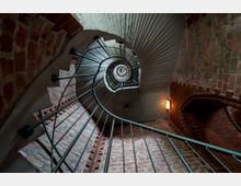 Spiralförmige Wendeltreppe mit Metallgeländer, umgeben von roten Ziegelwänden und einem Betondeckengewölbe. Ein Notausgangsschild mit Beleuchtung ist an der Wand angebracht.