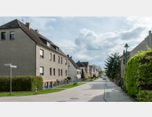 Straße mit Wohnhäusern in einer ruhigen Nachbarschaft, gesäumt von Grünflächen und Hecken. Die Häuser haben Schrägdächer und helle Fassaden, und die Straße ist gepflastert, mit Laternen auf beiden Seiten.