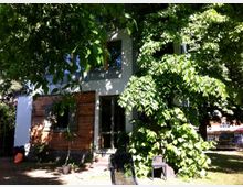 Zweistöckiges Wohnhaus mit heller Fassade und einem Abschnitt aus Holzverkleidung, umgeben von dichtem Grün und hohen Bäumen. Im Vordergrund befindet sich ein Garten mit Rasen sowie vereinzelten Gartenmöbeln.