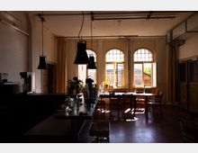 Innenraum eines Cafés oder Bistros mit Theke im Vordergrund, hängenden schwarzen Lampen und mehreren Holztischen mit Stühlen. Im Hintergrund lassen hohe Rundbogenfenster viel Tageslicht herein; an den Fensterbänken stehen kleine Pflanzen.