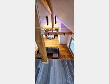 Innenansicht eines modernen Wohnbereichs mit offenem Dachstuhl aus Holz. Der Raum verfügt über eine Sitzecke mit Sofa, eine Küche mit Esstheke und Barhockern sowie einem Holzboden; sichtbare Holzbalken und große Dachfenster prägen das Design.