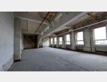 Ein leerer, verfallener Raum mit Betonboden und abgenutzten Wänden. Die Decke zeigt freiliegende Rohre, und an einer Seite befinden sich mehrere große Fenster mit Blick auf eine Außenlandschaft.