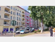 Straßenszene mit mehrstöckigen Wohnhäusern im Altbaustil, gelb und rosa gestrichen, mit Balkonen und großen Fenstern. Im Vordergrund eine Straße mit Autos, Fahrradfahrern und Fußgängern sowie Bäumen am rechten Bildrand.